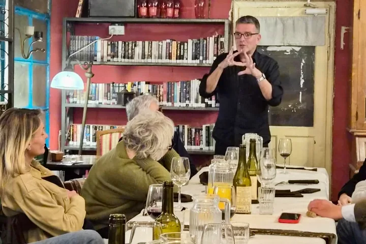A long table in a wine bar, people seated along it with glasses and jugs, Nir Tsuk standing at the head of it, talking with his hands.