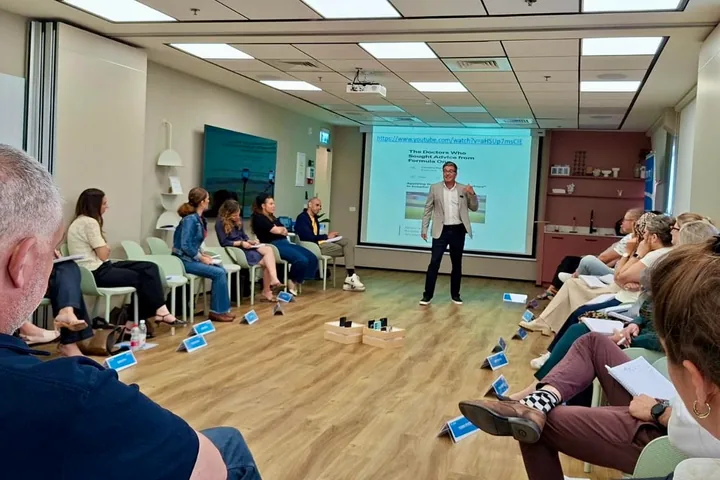 A seminar room: participants seated in a wide circle with notebooks, Nir Tsuk standing in the middle in front of a slide titled “The Doctors Who Sought Advice from Formula One”.