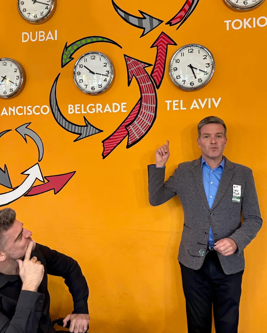 Nir Tsuk speaking in front of a wall of clocks labelled Dubai, San Francisco, Belgrade, Tel Aviv and Tokyo, one person listening